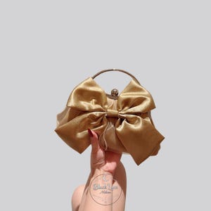 May include: A gold-colored clutch bag featuring a large, prominent satin bow on the front. The bag has a circular metal handle and a small, round metal clasp at the top. The surface of the bag has a smooth, reflective finish with a subtle sheen.