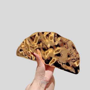 May include: A gold-colored, abstract-shaped decorative object with a textured surface. The object is held in a hand against a light gray background. The object's design features a swirling pattern of light and dark gold tones.