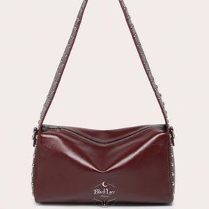 Può includere: Borsa a tracolla bordeaux con borchie argentate. La borsa rettangolare presenta una chiusura con zip e lunghe cinghie, anch'esse decorate con borchie. Il testo "Black Lace Nikita" è visibile in basso.
