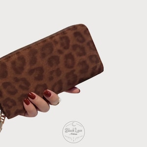 May include: A rectangular zip-around wallet featuring a brown leopard print pattern on a textured surface. The wallet has a gold-toned metal zipper pull and a matching gold-toned zipper track along the top and side edges of the accessory.