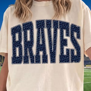 Op de afbeelding: Een crèmekleurig T-shirt met korte mouwen en het woord BRAVES in grote, dikke, donkerblauwe blokletters. De letters hebben een subtiel donkerblauw luipaardpatroon, wat zorgt voor een gestructureerde uitstraling op de lichte stof.