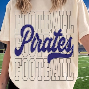 May include: A cream-colored short-sleeve t-shirt featuring a graphic design with the word FOOTBALL in a large outlined block font repeated at the top and bottom, with the word Pirates in a large dark blue cursive script across the center.