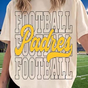 May include: A cream-colored short-sleeve t-shirt featuring a graphic print with the word FOOTBALL in black block letters repeated twice, with the word Padres written in a large, yellow, cursive script layered across the center of the design.