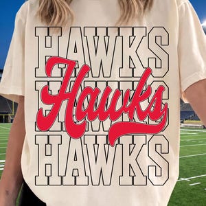 May include: A cream-colored short-sleeve t-shirt featuring a graphic design with the word HAWKS printed in a large black outlined block font on the top and bottom rows, with the word Hawks in a red cursive script font across the middle.