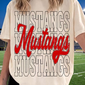 May include: A cream-colored short-sleeve t-shirt featuring the word MUSTANGS printed in a large black outlined block font on the top and bottom rows, with the word Mustangs in a bold red cursive script layered across the center.