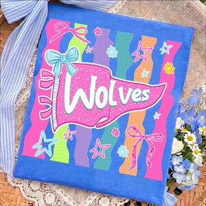 Peut inclure: Un article en tissu carré bleu présentant un fanion rose central avec le texte "WOLVES" en blanc. Le motif comprend des rayures ondulées verticales vertes, violettes et orange, décorées de nœuds, d'étoiles et de petites fleurs roses et bleues.