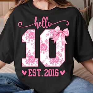 以下が含まれることがあります： 黒いTシャツ。ピンクの筆記体で「hello」の文字が、数字の「10」の上に書かれています。「10」はピンクの花柄で埋められ、ピンクのリボンで飾られています。「EST. 2016」の文字が下にあり、両側にピンクのハートがあります。