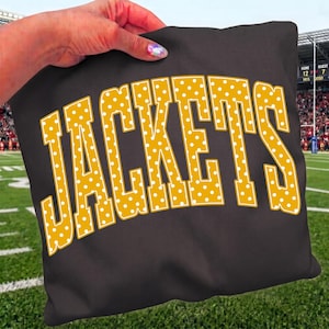 May include: A square black fabric pillow featuring the word JACKETS printed in a large, arched, gold-colored font. The letters are decorated with a repeating pattern of small white polka dots and outlined in a thin white border.