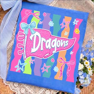 May include: A square blue fabric banner featuring a central pink pennant with white polka dots and the word Dragons in white script. The design includes colorful wavy vertical stripes, light blue bows, and various star and flower shapes.