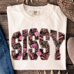 May include: Cream-colored t-shirt with the word "SISSY" in large, bold letters. The letters are filled with a pink and brown camouflage pattern. The shirt is folded on a wooden surface.