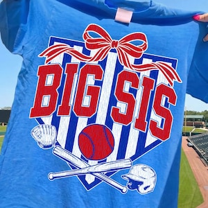 Puede incluir: Una camiseta azul con un diseño de béisbol. El diseño incluye las palabras "BIG SIS" en rojo, un lazo rojo, una pelota de béisbol, bates cruzados, un guante y un casco. La camiseta se muestra sobre un cielo azul.