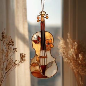 May include: Stained glass violin suncatcher with a butterfly and flower design. The violin features shades of amber, white, and brown. It hangs from a gold-colored chain, perfect for a window or wall decoration.