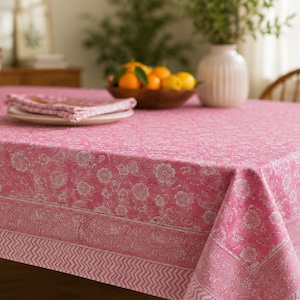 May include: A rectangular tablecloth featuring a vibrant pink base with a white floral pattern. The design includes a central section with intricate flowers, a wide border with a detailed botanical motif, and a bottom edge with a chevron pattern.