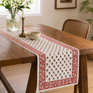 May include: A rectangular table runner featuring a cream-coloured base with a repeating pattern of small red and black floral motifs. The runner is bordered by a wide red band decorated with intricate white and red floral and vine designs.