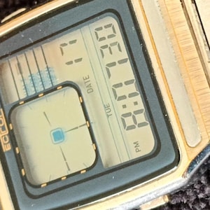 May include: Close-up of a gold-toned digital watch with a rectangular black face. The watch displays the time as 11:30 PM on Tuesday, with the date visible. The brand name "Ectron" is printed on the watch face.