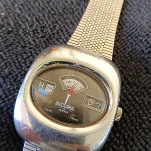 May include: A vintage Sicura watch with a silver-tone case and a mesh band. The watch face is dark with white and red details, including the date and day. The watch displays the date as 25/01/15.