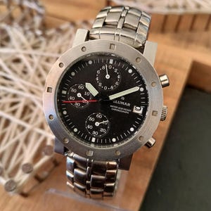 May include: A stainless steel wristwatch with a black face. The watch features multiple sub-dials, a date window, and the brand name "GLUMAR". The watch has a metal band and is displayed on a wooden surface.