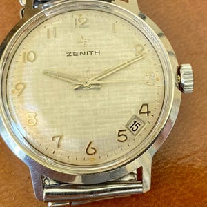 May include: Close-up of a vintage Zenith wristwatch with a silver-tone dial and hands. The watch features a date window at the 3 o'clock position and a silver-tone metal band. The watch face displays the word "ZENITH".