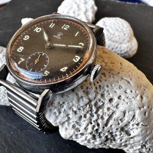 Könnte beinhalten: Vintage Armbanduhr mit schwarzem Zifferblatt, silberfarbenem Gehäuse und schwarzem Armband. Die Uhr hat eine kleine Sekundenscheibe und Leuchtzeiger. Die Uhr liegt auf einer hellen, strukturierten Oberfläche.