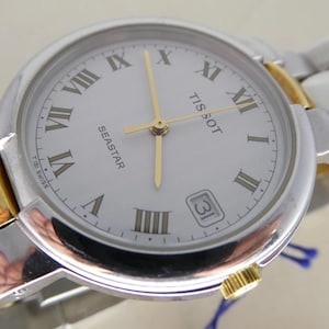 May include: Silver and gold-tone Tissot Seastar watch with a white face, gold hands, and Roman numeral hour markers. The watch features a date display at the 3 o'clock position and a silver and gold-tone band. The watch is a classic timepiece.