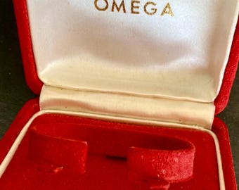 Può includere: Un cofanetto per orologio quadrato di colore rosso brillante con coperchio incernierato. L'interno è foderato in raso bianco sul coperchio superiore, con il logo e la scritta Omega in oro. La base è rivestita in velluto rosso con supporto.