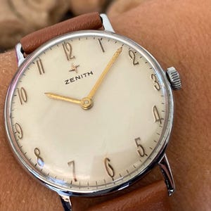 May include: A vintage Zenith wristwatch with a cream-colored face, gold hands, and a brown leather strap. The watch has a silver-colored case and displays the Zenith logo. The watch face has Arabic numerals.