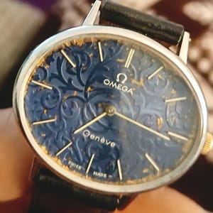 May include: An Omega Genève wristwatch with a dark blue face featuring an ornate pattern. The watch has gold-colored hands and hour markers, a silver-colored bezel, and a black leather band. The watch face also has the words "Omega", "Genève", "Swiss Made".