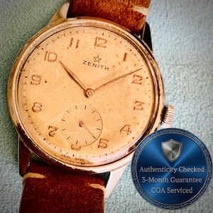 May include: A vintage Zenith wristwatch with a round, aged gold-toned dial, Arabic numerals, and a small sub-dial for seconds. The watch features a brown leather strap and a metallic crown. A circular blue badge displays Authenticity Checked text.