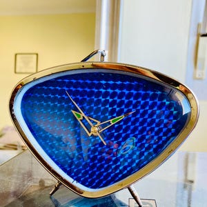 May include: A triangular-shaped analog clock with a metallic gold-toned frame and a vibrant blue textured dial. The clock face features a repeating geometric pattern and thin, pointed hands with small green accents at the tips of the hour and minute hands.