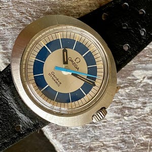 May include: An Omega Genève Dynamic watch with a round silver-tone case, a blue and silver dial featuring the Omega logo, and the text Genève DYNAMIC. The watch has black and blue hands and is attached to a black perforated leather watch strap.