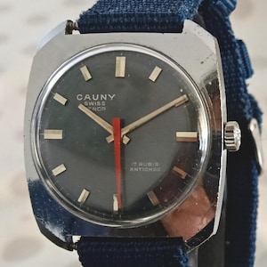 May include: A vintage silver wristwatch with a dark grey face and a navy blue fabric strap. The watch face has gold hands and markers, with the text "CAUNY SWISS NOR" and "17 RUBIS ANTICHOC". The second hand is red.