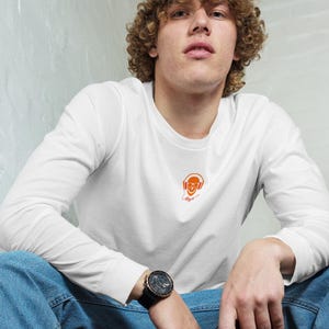 Puede incluir: Camiseta blanca de manga larga con un gráfico naranja de una calavera con auriculares. La camiseta se combina con vaqueros azules y una gorra de béisbol azul marino. También se ven un reloj negro y un teléfono negro.