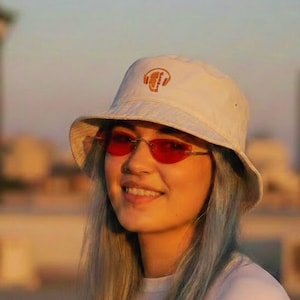 May include: A person wearing a white bucket hat with an orange logo, red-tinted sunglasses, and a white long-sleeve shirt. The person has long, light blue hair and is smiling. The background features a cityscape at sunset.