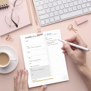 Puede incluir: Un planificador semanal con un dise&ntilde;o de cuadr&iacute;cula, sujeto con un clip dorado. El planificador incluye una lista de tareas y un horario semanal. Una mano sostiene un bol&iacute;grafo, marcando elementos de la lista. Un teclado y una taza de caf&eacute; est&aacute;n en el fondo.