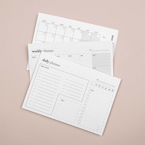 Puede incluir: Una pila de planificadores de papel blanco con dise&ntilde;o mensual, semanal y diario. El planificador diario incluye secciones para un horario, notas y una lista de tareas. El planificador semanal tiene un dise&ntilde;o de cuadr&iacute;cula. El planificador mensual es un calendario.