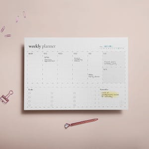 Puede incluir: Un planificador semanal blanco con un dise&ntilde;o de cuadr&iacute;cula, con el texto "weekly planner". El planificador incluye secciones para cada d&iacute;a de la semana, una lista de "To do" y una secci&oacute;n de "Remember". Un bol&iacute;grafo y clips de papel de oro rosa est&aacute;n en la superficie.