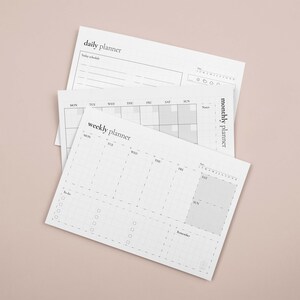 Puede incluir: Tres planificadores de papel blanco con el texto "daily planner", "weekly planner" y "monthly planner". Los planificadores tienen un dise&ntilde;o de cuadr&iacute;cula con espacios para notas y programaci&oacute;n. El fondo es de color rosa claro liso.