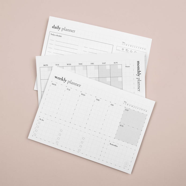 Puede incluir: Tres planificadores de papel blanco con el texto "daily planner", "weekly planner" y "monthly planner". Los planificadores tienen un dise&ntilde;o de cuadr&iacute;cula con espacios para notas y programaci&oacute;n. El fondo es de color rosa claro liso.