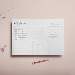 Puede incluir: Un planificador diario blanco con un patr&oacute;n de cuadr&iacute;cula, con un horario y una lista de tareas. El horario incluye horarios y actividades como "pasear al perro" y "cena familiar". Un bol&iacute;grafo y clips de papel de oro rosa est&aacute;n sobre un fondo rosa.