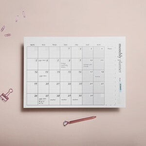 Puede incluir: Un planificador mensual blanco con un dise&ntilde;o de cuadr&iacute;cula, con fechas y espacio para notas. El planificador incluye el texto "monthly planner" y el mes de "Octubre". Un bol&iacute;grafo y clips de papel de oro rosa est&aacute;n sobre una superficie rosa claro.