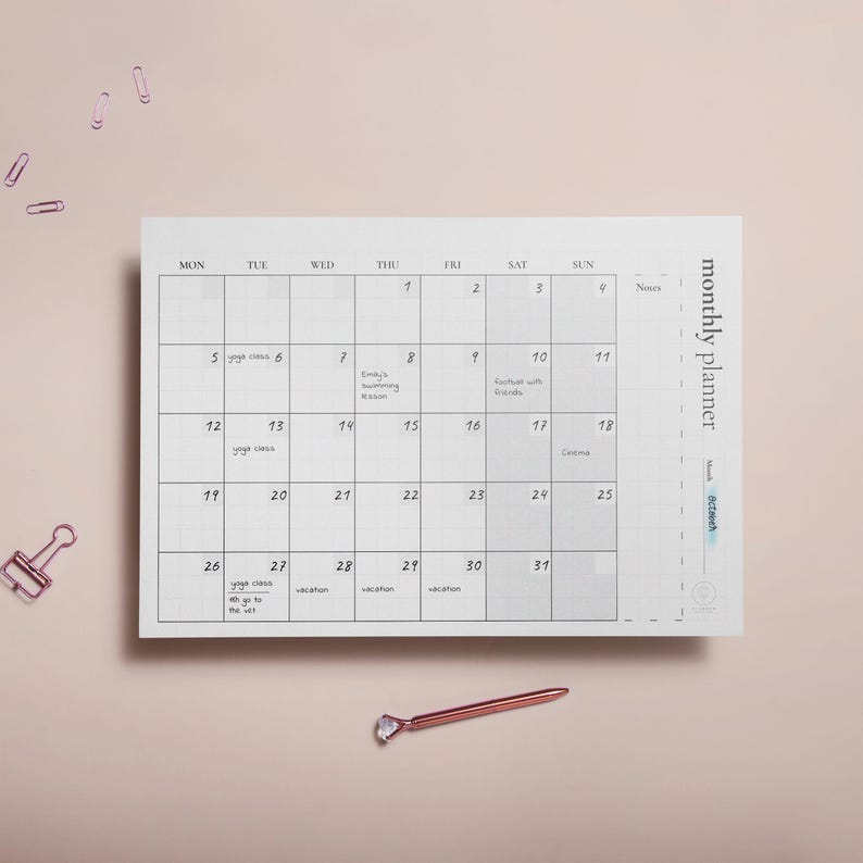 Puede incluir: Un planificador mensual blanco con un dise&ntilde;o de cuadr&iacute;cula, con fechas y espacio para notas. El planificador incluye el texto "monthly planner" y el mes de "Octubre". Un bol&iacute;grafo y clips de papel de oro rosa est&aacute;n sobre una superficie rosa claro.