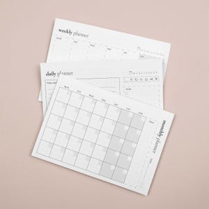 Puede incluir: Una pila de planificadores de papel blanco, que incluyen un planificador semanal, diario y mensual. Los planificadores tienen un dise&ntilde;o de cuadr&iacute;cula con secciones para fechas, notas y listas de tareas. Los textos "weekly planner", "daily planner" y "monthly planner" son visibles.