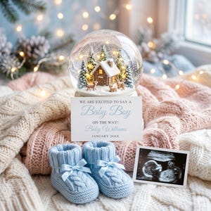 Peut inclure: Une boule à neige contenant une cabane miniature, des sapins et des cerfs, accompagnée d'une carte d'annonce blanche indiquant WE ARE EXCITED TO SAY A Baby Boy ON THE WAY! Baby Williams JANUARY 20XX, des chaussons en tricot bleu clair et une échographie.