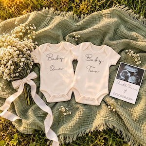 May include: Two cream-colored short-sleeve baby bodysuits with black text reading Baby One and Baby Two, accompanied by a bouquet of white baby's breath flowers with a cream ribbon and a rectangular ultrasound photo labeled Smith Twins October 202X.