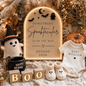 May include: An arched wooden sign with backlighting features the text SOMETHING SPOOKTACULAR IS ON THE WAY AUGUST 2026, a ghost, bats, and moon. Includes a crocheted ghost, a black cauldron labeled BABY BREWING, wooden BOO blocks, and a baby onesie.
