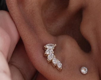 May include: A gold-toned ear stud featuring a curved arrangement of five marquise-cut clear stones, positioned alongside a single small round clear stone stud. The jewellery is worn on the lower earlobe, showcasing a delicate and bright metallic finish.