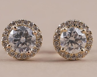 May include: A pair of round stud earrings crafted from yellow gold. Each earring features a large, central round-cut clear stone surrounded by a halo of smaller, pavé-set clear stones held in place by four prongs.