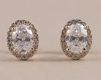 May include: A pair of stud earrings featuring a central oval-cut clear stone surrounded by a halo of small round clear stones set in a gold-toned metal frame. The earrings are symmetrical and display a classic prong setting design.