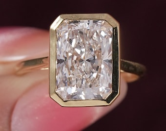 May include: A gold ring featuring a large, rectangular, radiant-cut clear gemstone set in a smooth, polished gold bezel setting. The band is thin and made of matching gold, providing a clean and focused look at the central stone.