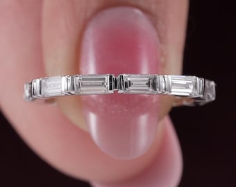 May include: A silver-toned eternity band ring featuring a continuous row of rectangular baguette-cut clear stones set in a channel setting. The metal band has a polished finish and the stones are arranged horizontally around the entire circumference.